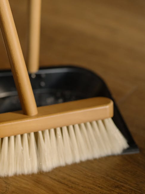 A light blue dustpan and a wooden-handled cleaning brush with beige bristles are hanging on a white wall, showing a clean and organized surface cleaning arrangement. The dustpan's smooth surface and t