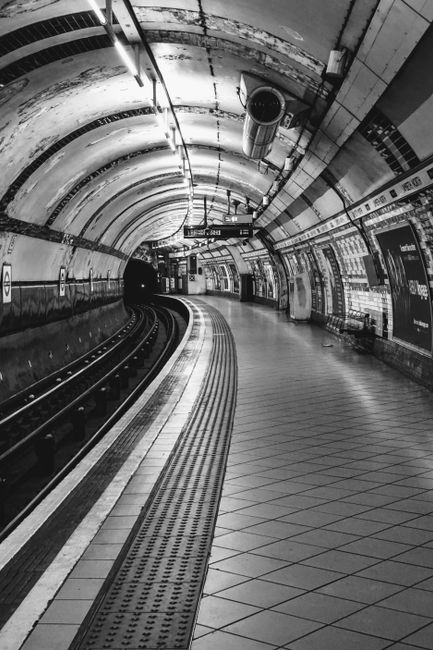 A low-angle view of the platform at Colliers Wood tube station, showcasing the textured tiled flooring with a central yellow safety line, flanked by advertisements on the curved wall, and a group of c