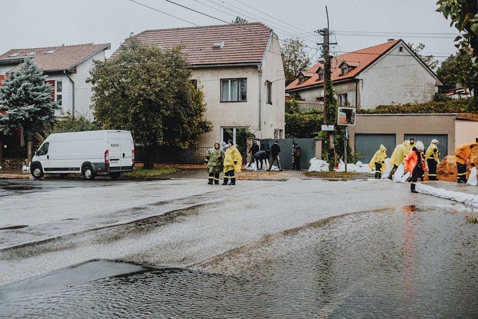 A residential street during an emergency flood response, with workers in yellow and orange waterproof suits and protective gear managing floodwaters that have accumulated on the cobblestone road. The