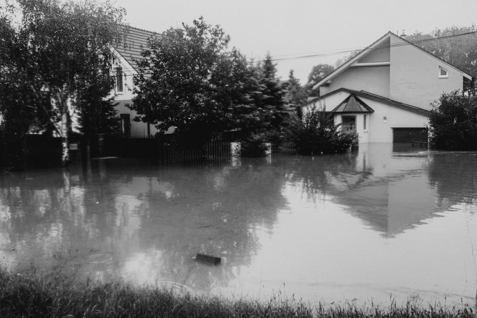 A residential street during an emergency flood response, with workers in yellow and orange waterproof suits and protective gear managing floodwaters that have accumulated on the cobblestone road. The